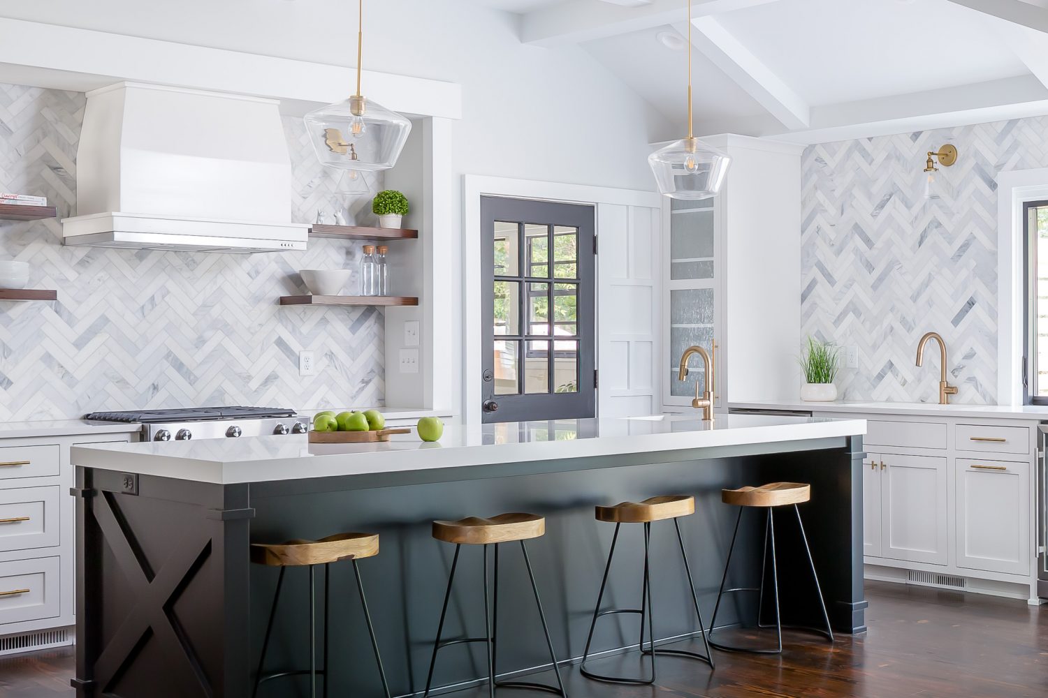 Modern white kitchen remodel with a large black island, marble herringbone backsplash, brass fixtures, glass pendant lighting, and warm wood bar stools in an open-concept layout.