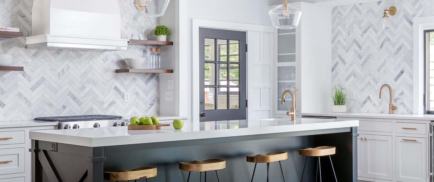 Glascock Modern white kitchen remodel with a large black island, marble herringbone backsplash, brass fixtures, glass pendant lighting, and warm wood bar stools in an open-concept layout.