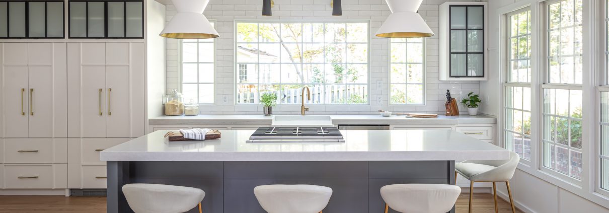 Bright, modern kitchen remodel featuring a large navy island with quartz countertops, white shaker cabinets, black framed frosted glass cabinet doors, brass hardware, exposed wood ceiling beams, oversized pendant lighting, and expansive windows that fill the space with natural light.