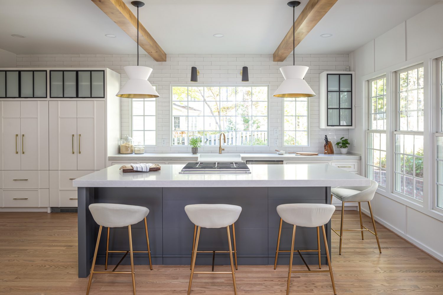 Bright, modern kitchen remodel featuring a large navy island with quartz countertops, white shaker cabinets, black framed frosted glass cabinet doors, brass hardware, exposed wood ceiling beams, oversized pendant lighting, and expansive windows that fill the space with natural light.