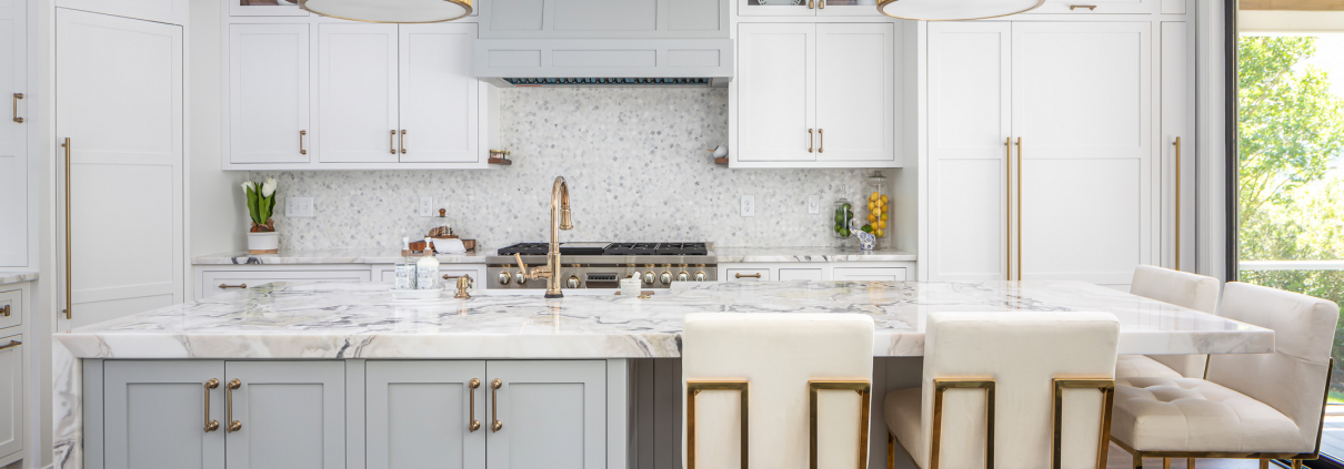 Luxury kitchen remodel with a large marble waterfall island, soft gray and white shaker cabinets, brass pendant lighting, integrated appliances, and elegant gold accents that create a bright, upscale gathering space.