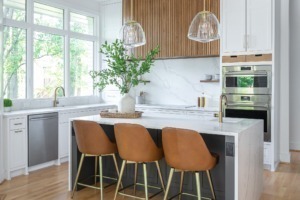 Modern kitchen remodel with a white waterfall island, black cabinetry painted sherwin williams black fox, warm wood slat custom vent hood with marble backsplash, brass fixtures, and leather barstools, creating a bright and contemporary space filled with natural light.