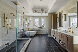 DSC03138 (Large) Bright primary bathroom with a freestanding soaking tub set within a bay window, glass-enclosed shower, double vanity, dark tile flooring, and makeup area with storage..