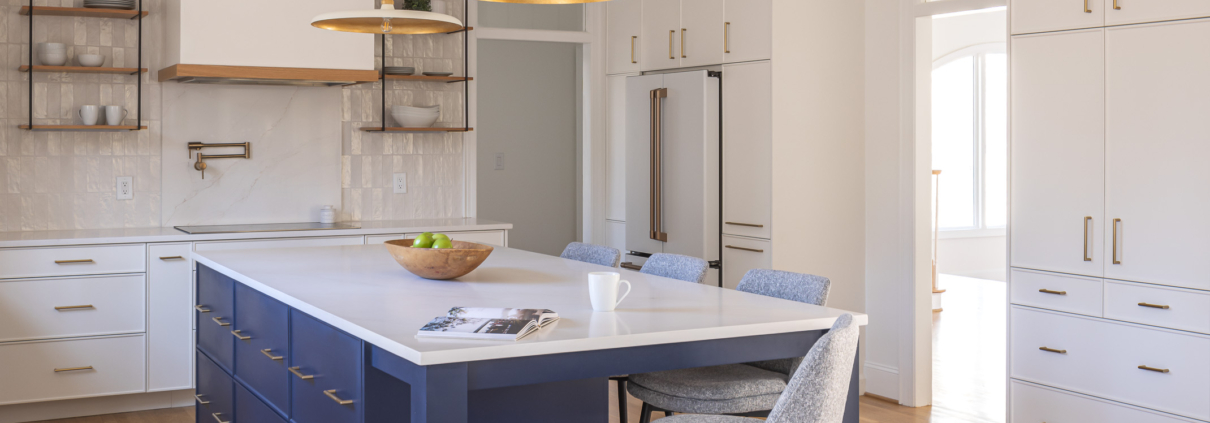 Bright transitional kitchen featuring a navy island, white cabinetry, quartz countertops, brass pendant lighting, warm wood shelving, and a layered tray ceiling detail with recessed lighting.