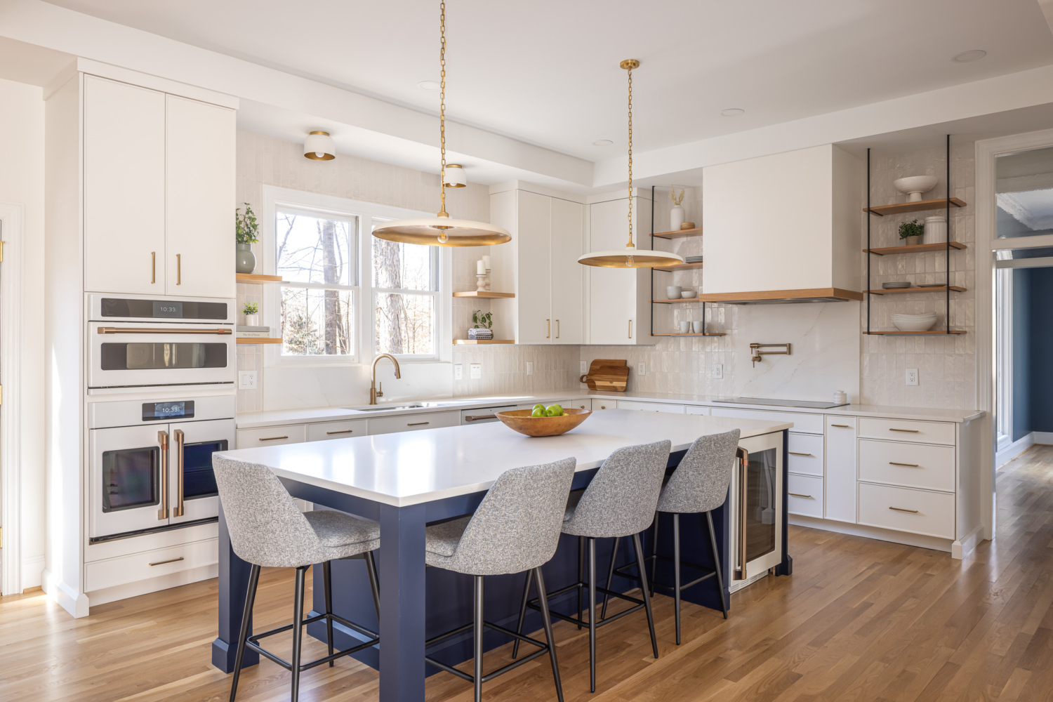 Bright transitional kitchen remodel in Raleigh NC with white custom cabinetry, navy blue island, brass pendant lighting, open shelving, and warm hardwood floors in an airy open-concept design.