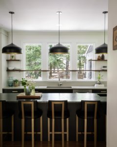 a_12 Modern kitchen with a dark waterfall island, black and brass pendant lights, brass-accented bar stools, floating wood shelves, and large windows above the sink.