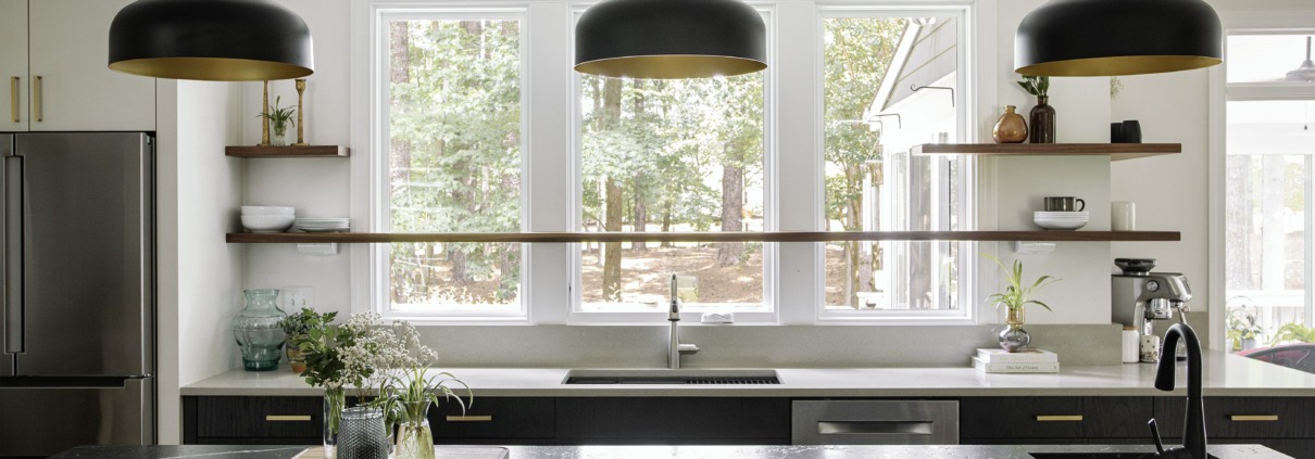 Modern kitchen with a black waterfall island, matte black pendant lights, white cabinetry, floating wood shelves, and large windows for natural light.