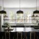 Modern kitchen with a black waterfall island, matte black pendant lights, white cabinetry, floating wood shelves, and large windows for natural light.