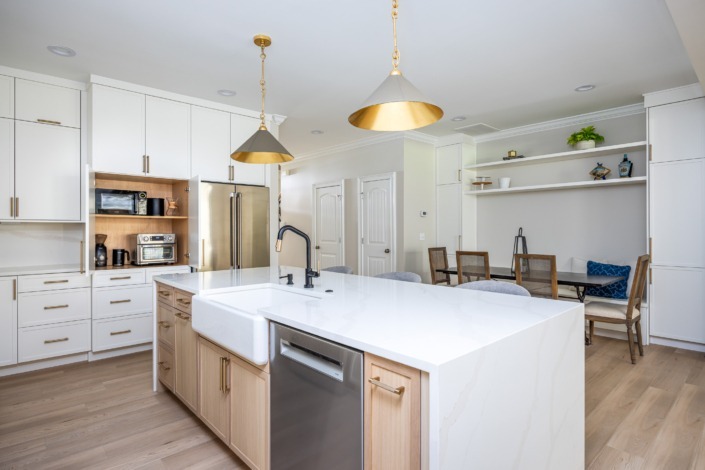 Bright transitional kitchen remodel in Raleigh NC with a large waterfall island, farmhouse sink, white shaker cabinetry, brass pendant lighting, and warm wood accents, featuring hidden appliance storage for a clean and organized design.