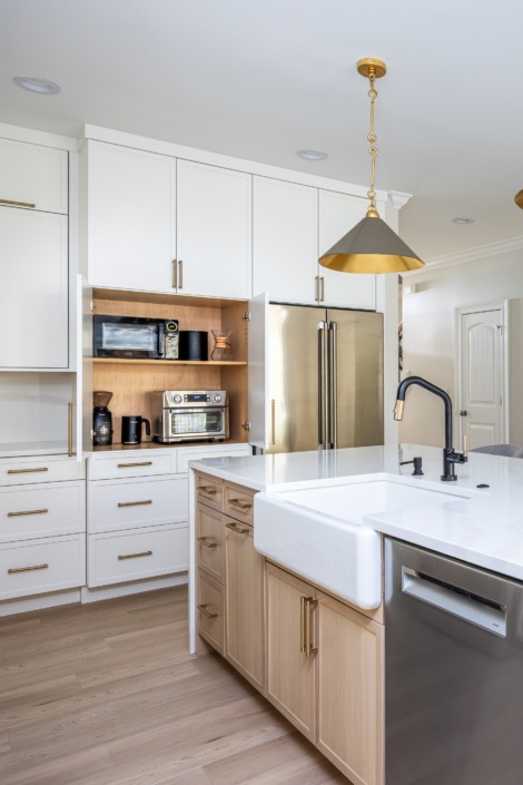 Bright transitional kitchen remodel in Raleigh NC with a large waterfall island, farmhouse sink, white shaker cabinetry, brass pendant lighting, and warm wood accents, featuring hidden appliance storage for a clean and organized design.