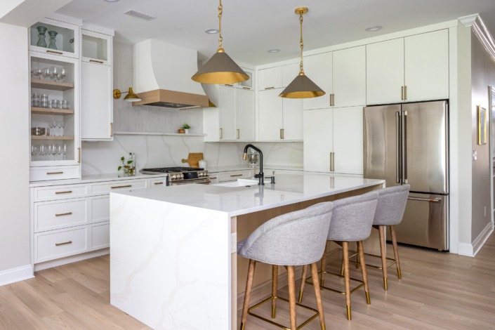 Modern white kitchen remodel in Raleigh NC with custom cabinetry, brass hardware, textured tile backsplash, wood range hood, waterfall island, and warm contemporary lighting.