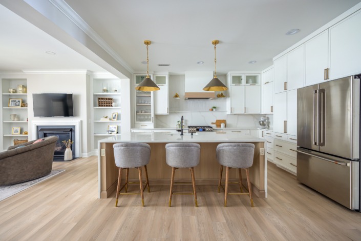 Bright transitional kitchen with white custom cabinetry, warm wood island accents, brass pendant lighting, waterfall countertops, and open-concept luxury design.