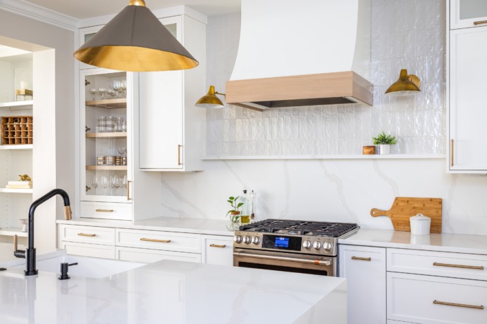 Bright white transitional kitchen remodel in Raleigh NC with waterfall island, brass pendant lighting, custom range hood, glass-front cabinetry, and warm wood accents for a timeless luxury design.