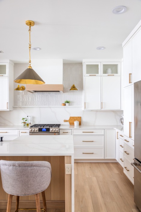 Modern white kitchen remodel in Raleigh NC with custom cabinetry, brass hardware, textured tile backsplash, wood range hood, waterfall island, and warm contemporary lighting.