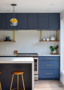 clearcut-7813-netherlands-10 Close-up of a modern navy kitchen with flat-panel cabinetry, floating wood shelves with brass supports, quartz backsplash, globe pendant lighting, and a sleek waterfall island that creates a bold contemporary aesthetic.
