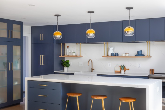 Bold contemporary kitchen remodel in Cary NC featuring deep navy flat-panel cabinetry, a large white waterfall island, brass hardware, floating wood shelves, globe pendant lighting, and warm wood barstools for a sleek and sophisticated modern design.
