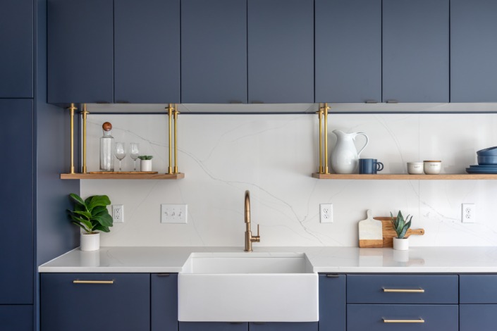 Modern navy kitchen design with sleek flat-panel cabinetry, brass hardware, floating wood shelves, quartz backsplash, and a white farmhouse sink that creates a clean and sophisticated contemporary space.