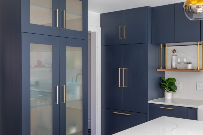 Close-up of bold navy kitchen cabinetry with brass hardware, frosted glass cabinet doors, floating wood shelves with brass supports, and sleek quartz countertops that create a refined modern kitchen aesthetic.