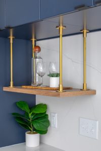 clearcut-7813-netherlands-15 Close-up of a custom floating wood shelf with brass hanging supports, with deep navy cabinetry and a white quartz backsplash, modern glassware and greenery.