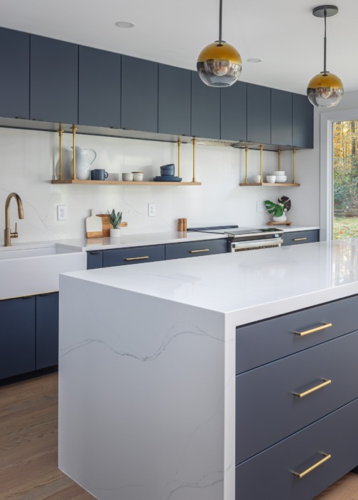 Close-up of a bold contemporary kitchen featuring deep navy flat-panel cabinetry, a dramatic white waterfall quartz island with storage on the seating side, brass hardware, and floating wood shelves for a sleek Scandinavian design.