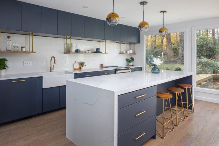 Close-up of a bold contemporary kitchen featuring deep navy flat-panel cabinetry, a dramatic white waterfall quartz island with storage on the seating side, brass hardware, floating wood shelves, and globe pendant lighting for a sleek modern aesthetic.