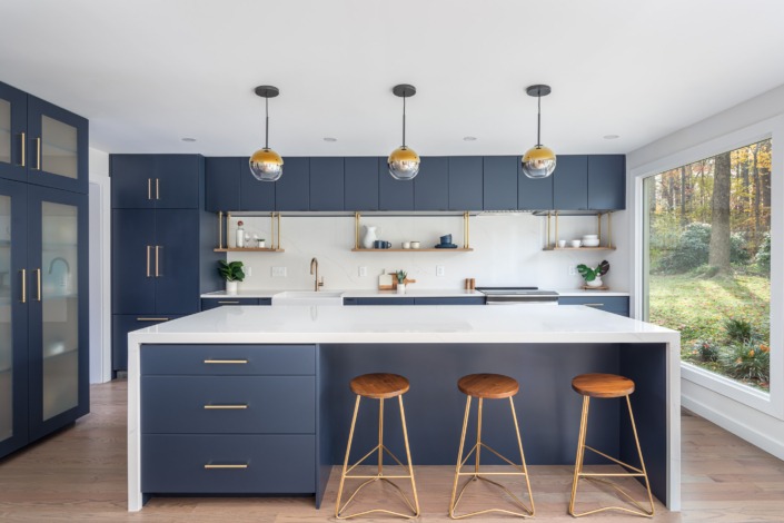 Bold modern kitchen remodel featuring deep navy cabinetry, a large white waterfall island, brass hardware, floating wood shelves, globe pendant lighting, and expansive picture windows that create a sleek and sophisticated open-concept space.