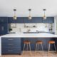 A bold modern kitchen with deep navy cabinets, a large white waterfall island, brass hardware, floating wood shelves, globe pendant lights, and large picture windows.