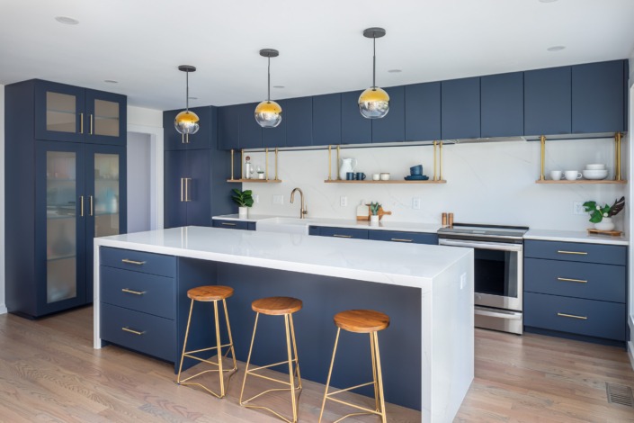 Bold contemporary kitchen remodel in Cary NC featuring deep navy flat-panel cabinetry, a large white waterfall island, brass hardware, floating wood shelves, globe pendant lighting, and warm wood barstools for a sleek and sophisticated modern design.