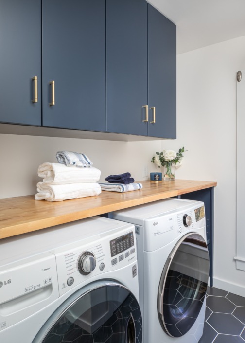 Modern laundry room design featuring deep navy custom cabinetry with brass hardware, a warm wood countertop, front-loading washer and dryer, and dark hexagon tile flooring for a clean and sophisticated utility space.