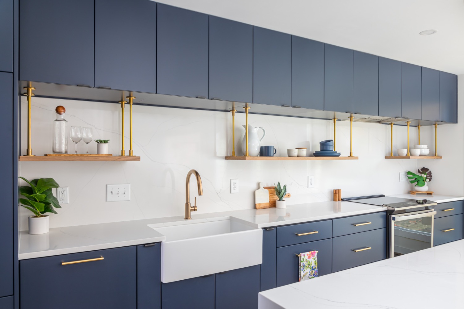 Modern navy kitchen remodel with sleek flat-panel cabinetry, a white farmhouse sink, quartz countertops and backsplash, floating wood shelves with brass supports, and warm brass fixtures that create a clean and sophisticated contemporary design.