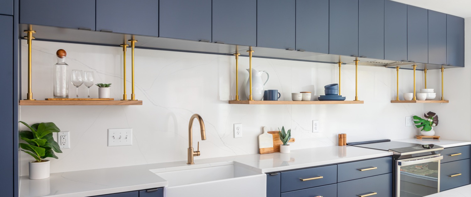 clearcut-7813-netherlands-8 Modern navy kitchen remodel with flat-panel cabinetry, a white apron front farmhouse sink, seamless quartz countertop and backsplash, floating wood shelves with hanging brass supports, and brass fixtures.