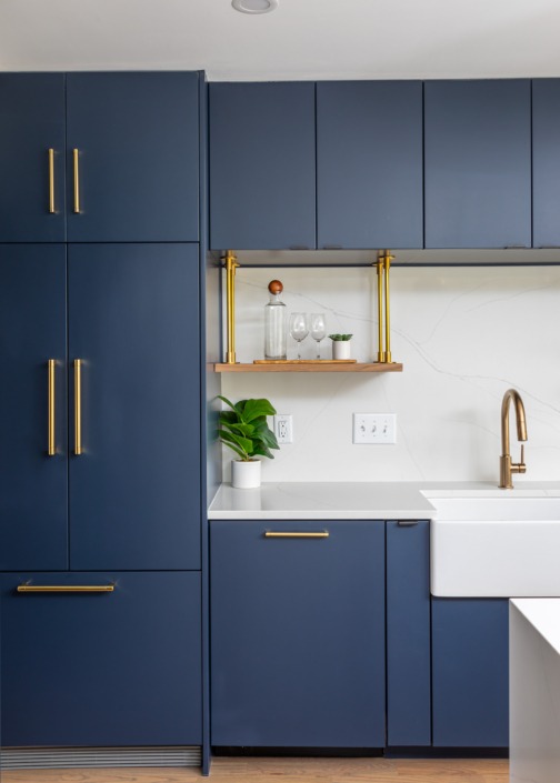 Close-up of a modern navy kitchen featuring flat-panel cabinetry, floating wood shelves with brass supports, quartz backsplash, globe pendant lighting, and a sleek waterfall island that creates a bold contemporary aesthetic.