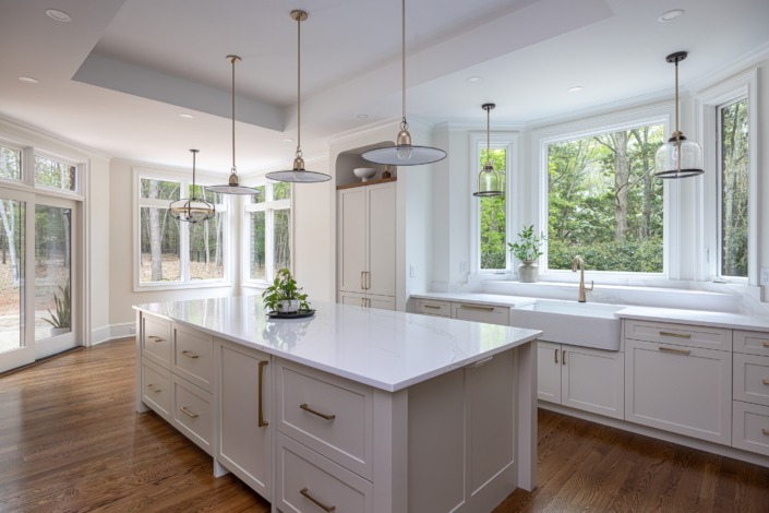 Elegant white kitchen remodel in Raleigh NC with a spacious quartz island, custom shaker cabinetry, brass hardware, layered pendant lighting, and a large window over the farmhouse sink that fills the space with natural light and warmth.