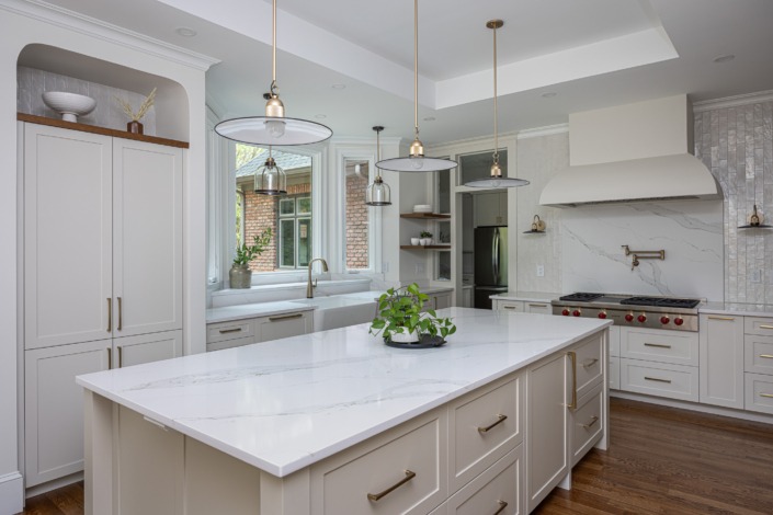 Elegant white kitchen remodel in Raleigh NC with a spacious quartz island, custom shaker cabinetry, brass hardware, layered pendant lighting, and a large window over the farmhouse sink that fills the space with natural light and warmth.