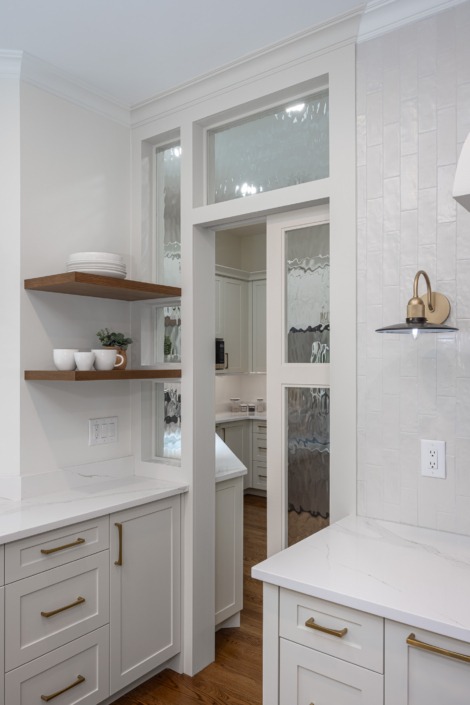 Custom white kitchen cabinetry with warm brass hardware, floating wood shelves, textured vertical tile backsplash, and decorative glass doors leading into the scullery for added storage, prep space, and timeless architectural charm.