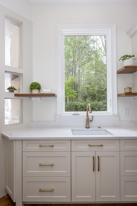 Pantry sink area featuring a large picture window with wooded views, a farmhouse apron-front sink, warm brass faucet and hardware, and elegant pendant lighting that fills the space with natural light and timeless charm. The scullery allows for added storage, prep space, and timeless architectural charm.