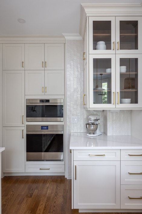 Bright white kitchen remodel with custom inset cabinetry, a textured vertical tile backsplash, glass-front upper cabinets, integrated double wall ovens, and warm brass hardware that adds timeless elegance and refined functionality to the space.