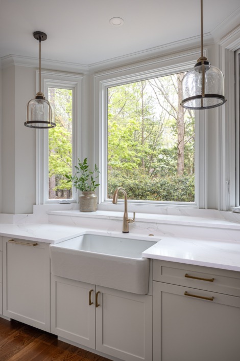 Bright kitchen sink area featuring a large picture window with wooded views, a farmhouse apron-front sink, warm brass faucet and hardware, and elegant pendant lighting that fills the space with natural light and timeless charm.