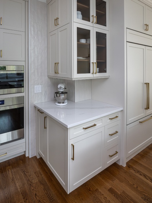 Custom kitchen storage detail featuring a built-in pull-out organizer beside the wall oven, white shaker cabinetry with brass hardware, under-cabinet lighting, and a dedicated countertop appliance station for added functionality and convenience.