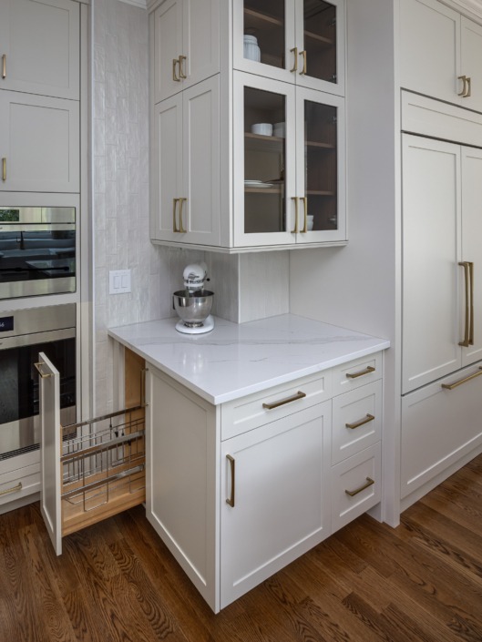Custom kitchen storage detail featuring a built-in pull-out organizer beside the wall oven, white shaker cabinetry with brass hardware, under-cabinet lighting, and a dedicated countertop appliance station for added functionality and convenience.
