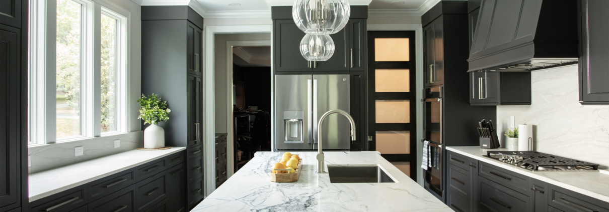 moody Wake Forest kitchen remodel with dark black cabinets painted in Sherwin Williams Iron Ore with white quartz on cabinets, island, and backsplash. A door well lit is at the back of the picture leading to the pantry.