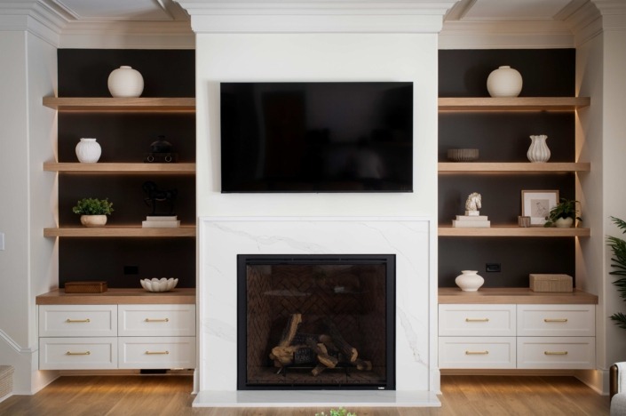 Wakefield_IMG_0014 Basement lounge featuring a modern fireplace with mounted TV, flanked by custom built-in cabinetry and warm wood floating shelves.