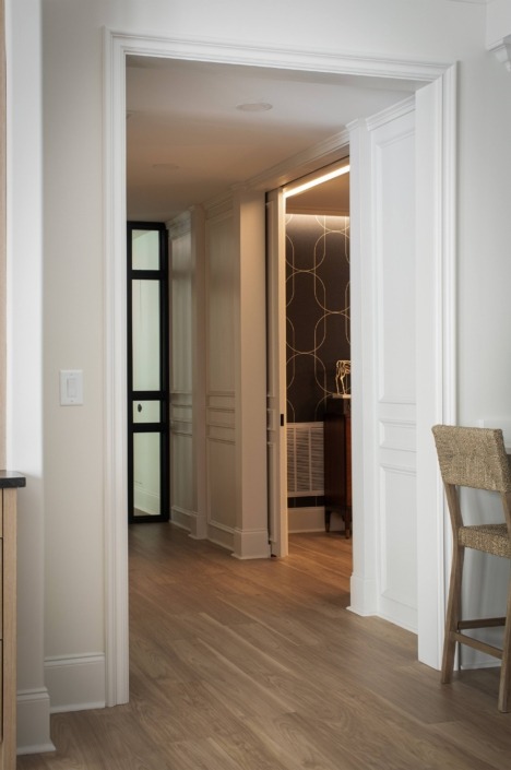 Wakefield_IMG_0017 Hallway leading to a basement lounge with hidden paneled doors and a home gym enclosed by black-framed glass doors.